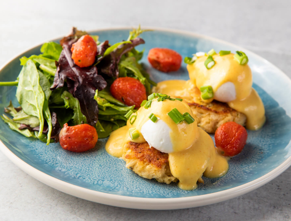 Two extra-large poached eggs served on two crab cakes, blistered grape tomatoes, scallions, cayenne citrus hollandaise, lemon vinaigrette salad instead of grits or house potatoes